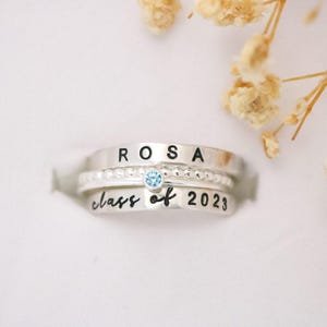 May include: Three silver rings stacked together. The top ring has the name "ROSA" stamped on it. The middle ring has small beads and a light blue gemstone. The bottom ring says "class of 2023".