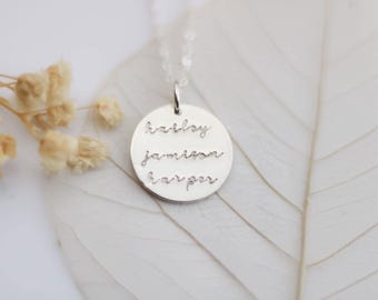 May include: A silver pendant necklace with a round charm engraved with the names "karsley", "jamison", and "harper" in a cursive font. The necklace is displayed on a white leaf with dried flowers in the background.