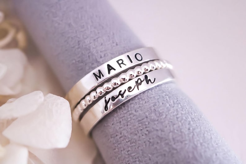 A silver ring with the name "MARIO" engraved on it, placed on a textured gray surface.