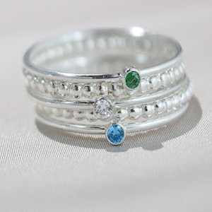 May include: A stack of three silver rings with beaded detailing. Each ring features a different colored gemstone: green, clear, and blue. The rings are displayed on a light-colored, textured surface.