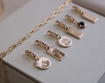 May include: Gold-colored jewelry charms and a chain are displayed on a light gray surface. The charms feature various designs, including a rainbow, a flower, and a gemstone. The chain has a link design.