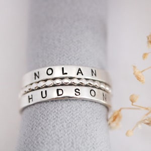 May include: Three silver rings stacked together, each with a different design. The top ring reads "NOLAN", the middle ring has a beaded design, and the bottom ring reads "HUDSON". The rings are displayed on a gray surface.