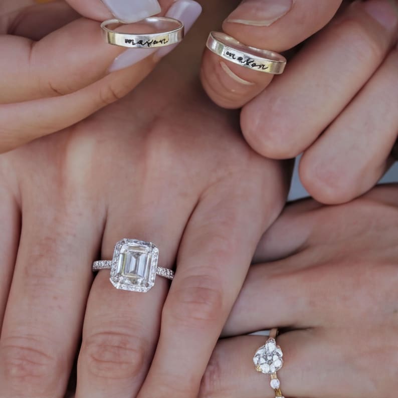 A close-up of a person's hands wearing various rings, including a large diamond ring and two rings with personalized messages engraved on them.