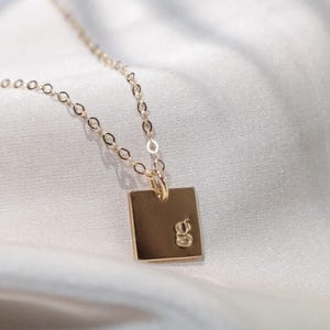 May include: A gold-colored necklace with a square pendant. The pendant has a small, engraved letter "g" and hangs from a delicate chain. The necklace is displayed on a white, silky fabric.