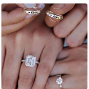 May include: Close-up of hands holding two silver rings engraved with the name "mason". A large square-cut diamond engagement ring and a smaller ring with a floral design are also visible. The rings are likely for a wedding or engagement.