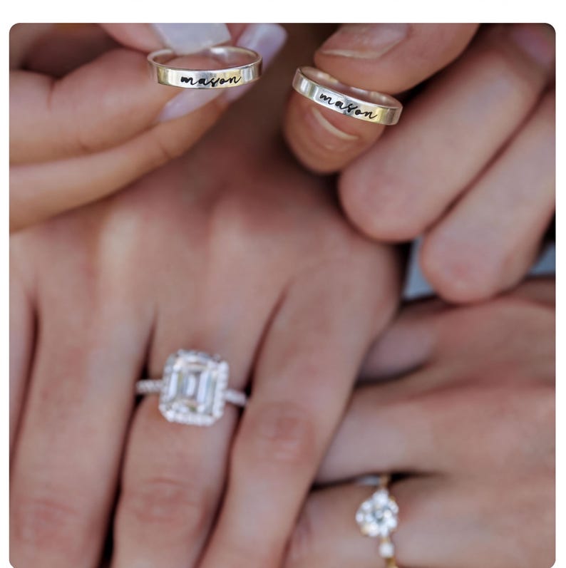 May include: Close-up of hands holding two silver rings engraved with the name "mason". A large square-cut diamond engagement ring and a smaller ring with a floral design are also visible. The rings are likely for a wedding or engagement.