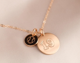 May include: A gold-colored necklace with two pendants. One pendant is a small, round charm with a baseball design. The other is a larger, round charm with the number 19 engraved on it. The necklace is on a light pink background.