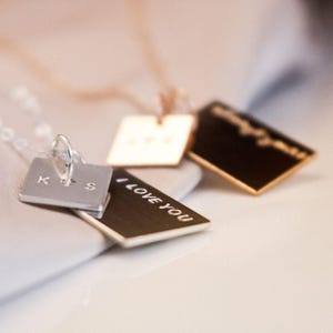 May include: Close-up of several pendant necklaces. One silver square pendant has the initials "K+S". Another pendant is gold, and the third is black with gold text that reads "I LOVE YOU". The necklaces are on a white surface.