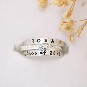May include: Three silver rings stacked together. The top ring has the name "ROSA" stamped on it. The middle ring has small beaded details and a light blue gemstone. The bottom ring says "class of 2023".