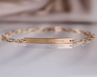 May include: A gold-colored bracelet with a chain link design. The bracelet features a rectangular bar in the center with the name "emma" engraved in cursive. The bracelet is displayed on a reflective surface.