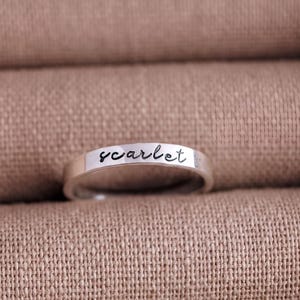 A silver ring with the word "scarlet" engraved on it, resting on a textured beige fabric.