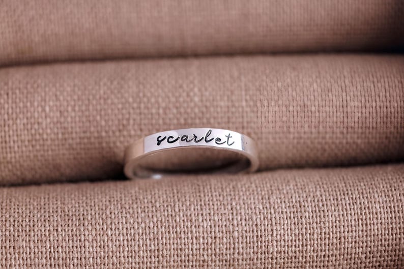 A silver ring with the word "scarlet" engraved on it, resting on a textured beige fabric.
