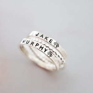 May include: Three silver stackable rings. The top ring reads "JAKE" with a paw print, the middle ring reads "MURPHY" with a paw print, and the bottom ring has a textured design. The rings are on a light background.