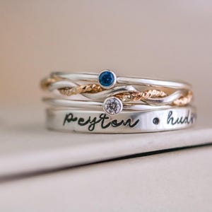 May include: A stack of silver rings with various designs. One ring features a twisted gold band, while others have small gemstones. The bottom ring is engraved with the name "Peyton" and the word "hudson".