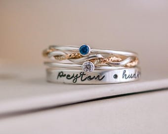 May include: A stack of silver rings with various designs. One ring features a twisted gold band, while others have small gemstones. The bottom ring is engraved with the name "Peyton" and the word "hudson".