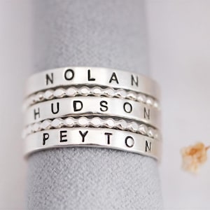 A silver ring with the names "Nolan", "Hudson", "Peyton" engraved on it, placed on a gray surface.