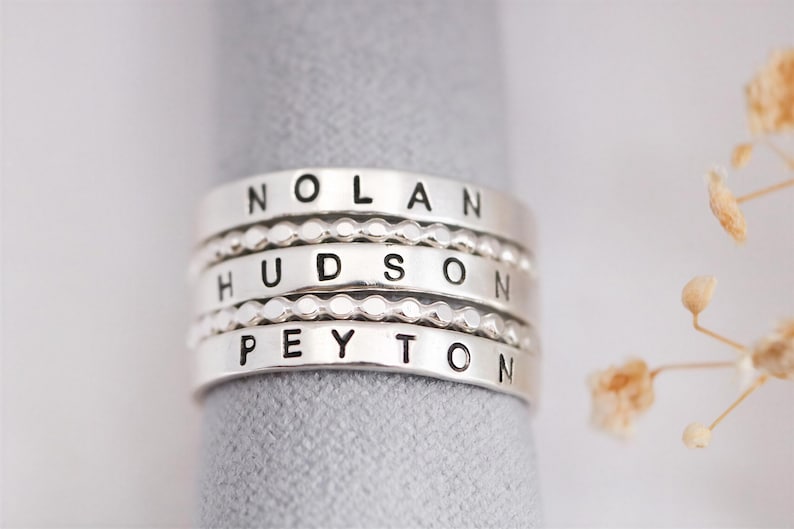 A silver ring with the names "Nolan", "Hudson", "Peyton" engraved on it, placed on a gray surface.
