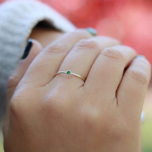 May include: A delicate silver ring with a small, round, green gemstone set in the center. The ring is simple and elegant, perfect for everyday wear. The ring is shown on a person's finger.