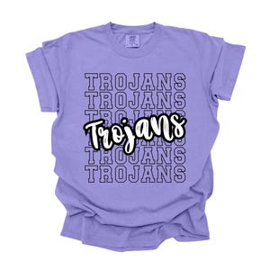 May include: Purple t-shirt with the word "Trojans" repeated in a stacked pattern in black outlined text. The word "Trojans" is also written in a larger, white outlined font in the center of the shirt.