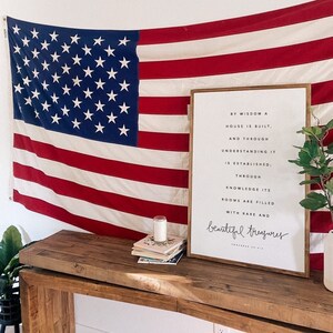 Pode incluir: Uma impressão emoldurada com fundo branco e texto preto que diz "Pela sabedoria se constrói uma casa, e pelo entendimento se estabelece; pelo conhecimento seus quartos são preenchidos com tesouros raros e preciosos." A impressão está pendurada em uma parede branca com uma bandeira americana drapeada atrás. Uma mesa de madeira com uma pilha de livros e uma vela fica na frente da impressão.