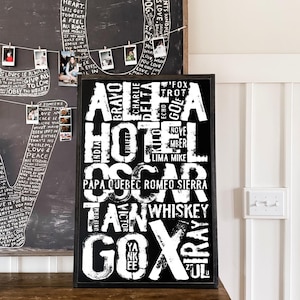 May include: A black and white word art print with a large white "X" in the center. The words surrounding the "X" include "Alta", "Hotel", "Oscar", "Tan", "Whiskey", "Gox", and more. The print is framed in a black frame and is hanging on a wall above a wooden table.