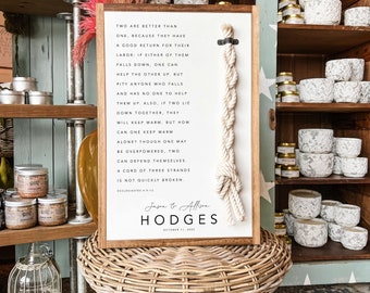 May include: A white wooden sign with a brown frame and a rope knot hanging from it. The sign has a quote about the strength of unity and a wedding announcement for Jason & Allison Hodges, October 11, 2022.