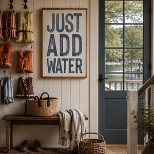 May include: A rectangular wall sign with a wooden frame featuring the dark blue text "JUST ADD WATER" in a bold, sans-serif font. The sign is displayed on a white paneled wall above a wooden bench, surrounded by hanging life vests and baskets.