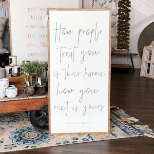 May include: A white wooden sign with a black handwritten quote: "How people treat you is their karma; how you react is yours - Wayne Dyer".