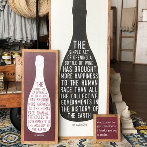 May include: Three wooden signs with a wine theme. The largest sign features a black silhouette of a wine bottle with the text "The simple act of opening a bottle of wine has brought more happiness to the human race than all the collective governments in the history of the earth. Jim Harrison". The medium sign features a red silhouette of a wine bottle with the same text. The smallest sign is red and says "Wine is good for your complexion, it breaks you out in smiles."