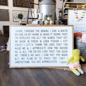 Könnte beinhalten: Ein weißes Holzschild mit schwarzem Text, der lautet: "'Cause through the drama I can always depend on my mama & when it seems that I'm hopeless you say the words that get me back in focus & even though I act crazy I gotta thank the Lord that you made me & I appreciate how you raised me & all the extra love that you gave me. There's no way I can pay you back but my plan is to show you that I understand. You are appreciated - tupac"