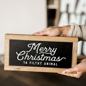 May include: A wooden sign with a black chalkboard background. The sign says "Merry Christmas Ya Filthy Animal" in white cursive lettering.
