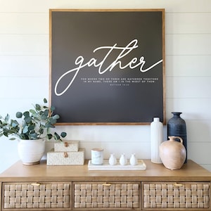 以下が含まれることがあります： 白い筆記体で「gather」と書かれた、濃い茶色の木製の額縁付きの看板。看板には、白いテキストで聖書の言葉「私の名において二人または三人が集まっているところに、私もその中にいる」も書かれている。看板は、籐製の引き出しが付いた木製のドレッサーの上、白い羽目板の壁に掛けられている。