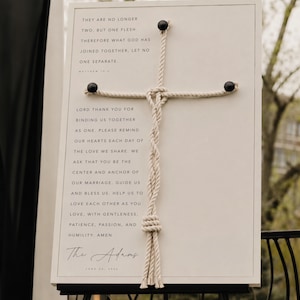 May include: A framed wedding sign with a cream-colored background and a rope cross. The sign includes the text "They are no longer two, but one flesh..." and a prayer. The sign is signed "The Adams" with the date June 20, 2024.