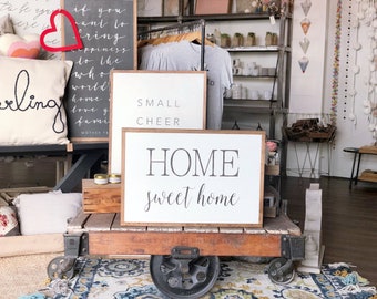May include: A wooden sign with the text "HOME sweet home" in black lettering on a white background. The sign is sitting on a wooden cart with wheels. There are other signs in the background, including one that says "SMALL CHEER".