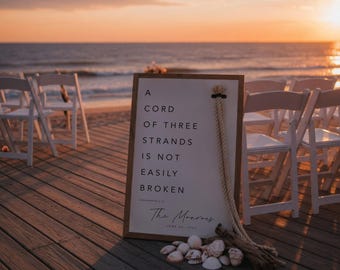 May include: A framed sign with the text "A cord of three strands is not easily broken" stands on a wooden deck near the beach. A rope and seashells are attached to the sign. White chairs are arranged on the beach, with the ocean and sunset in the background.