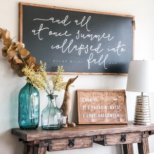 May include: A wooden console table with two drawers, decorated with a black chalkboard sign with the quote "and all at once summer collapsed into fall..." by Oscar Wilde, a wooden sign with the text "Look it, it's freakin bats, I love Halloween", two blue glass vases with yellow flowers, a wooden duck figurine, and a silver lamp with a white lampshade.
