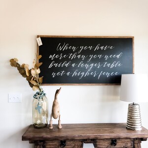 May include: A black chalkboard sign with a white wooden frame hangs on a white wall. The sign reads "When you have more than you need build a longer table not a higher fence".