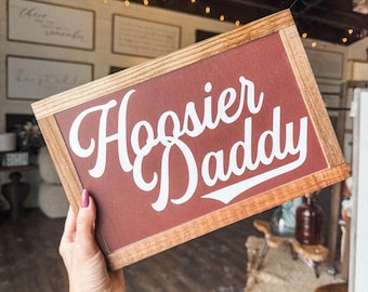 Puede incluir: Un letrero de madera con fondo rojo y letras blancas que dice "Hoosier Daddy". El letrero está en la mano de una persona.