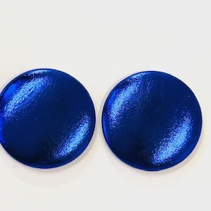 Puede incluir: Dos pendientes redondos de color azul metálico con una superficie texturizada. Los pendientes son de un tono azul vibrante y parecen reflejar la luz. Los pendientes están sobre un fondo blanco.