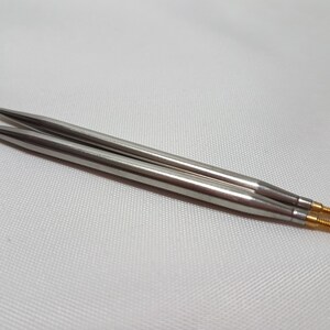 Puede incluir: Dos agujas de tejer plateadas con puntas doradas. Las agujas est&aacute;n sobre una superficie blanca.