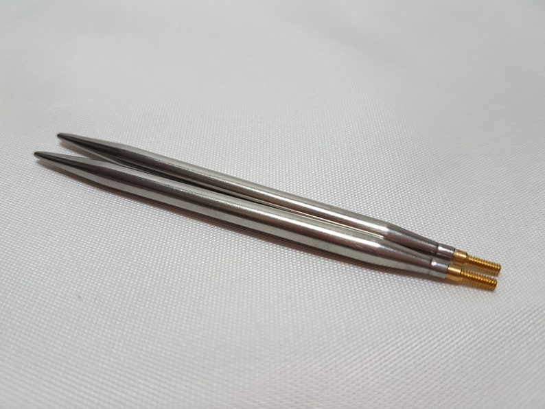 Puede incluir: Dos agujas de tejer plateadas con puntas doradas. Las agujas est&aacute;n sobre una superficie blanca.