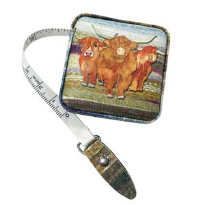 May include: A retractable measuring tape with a decorative case featuring a watercolor image of three Highland cattle. The tape extends to 150 cm and is white with black markings. The case is rectangular with a metal clasp.