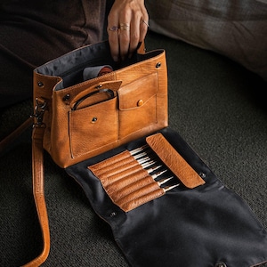 Può includere: Borsa da lavoro a maglia in pelle marrone chiaro con tracolla, aperta per rivelare ferri da maglia e accessori. La borsa ha una tasca frontale e una patta con chiusura a scatto. Gli aghi sono in una custodia di pelle marrone.