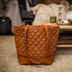 Può includere: Una borsa tote in pelle marrone con un design trapuntato e la parola "MUUD" impressa sul davanti.