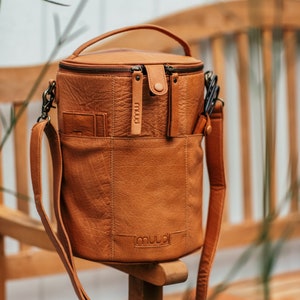 May include: A brown leather cylindrical bag with a zip closure and a shoulder strap. The bag has the word "muud" embossed on the front.