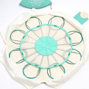 May include: A white canvas bag with a circular opening, revealing a set of circular knitting needles organised in a radial pattern. The needles are held in place by a clear plastic mesh. The bag has a teal green lining and a teal green zippered pouch with a white circle.