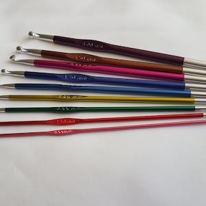 Könnte beinhalten: Ein Satz von zehn bunten Stricknadeln mit silbernen Spitzen. Die Nadeln sind nach Größe geordnet, wobei die kleinste Nadel oben und die größte Nadel unten liegt. Die Nadeln sind mit ihrer Größe in Millimetern beschriftet.