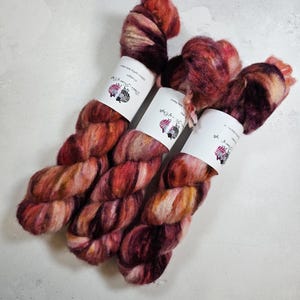 May include: Three skeins of hand-dyed yarn in shades of red, orange, and brown. The yarn is a blend of silk and mohair, and it has a soft, fuzzy texture. The skeins are labeled "Zebra & Quill" and "Hand Dyed Yarn".