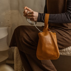 May include: A person wearing a brown sleeveless dress and black long-sleeved shirt is sitting on a white fluffy surface. They are holding a brown leather tote bag with a single handle. The person is knitting with white yarn.