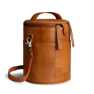 Può includere: Un portabottiglie cilindrico in pelle marrone, con un manico superiore e una tracolla staccabile. La borsa ha una parte superiore con cerniera, una tasca laterale e un'etichetta in pelle con la parola "muud".